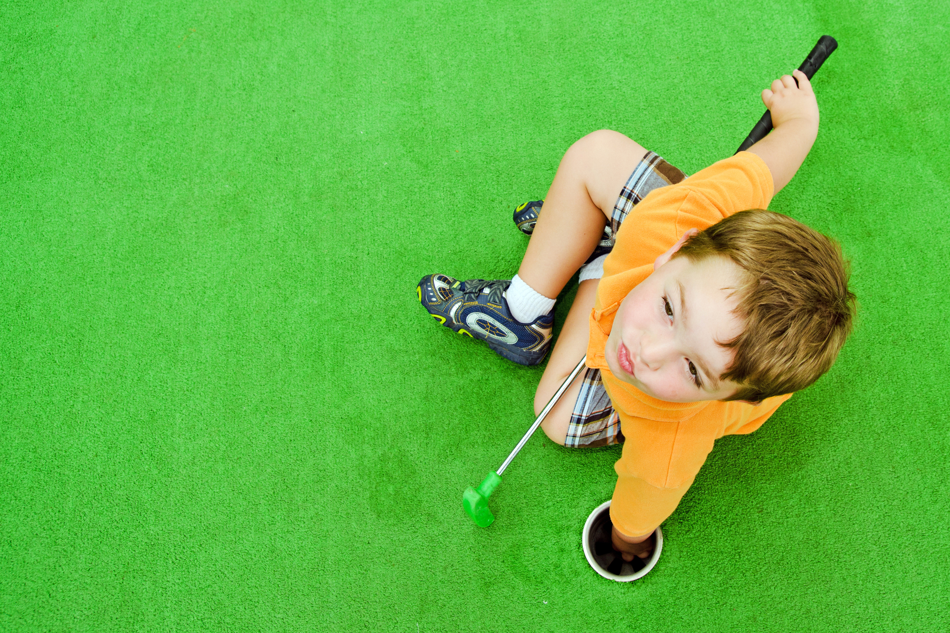 Kid reaching for golf ball in hole and sitting on artificial grass - Orion Turf and Landscape